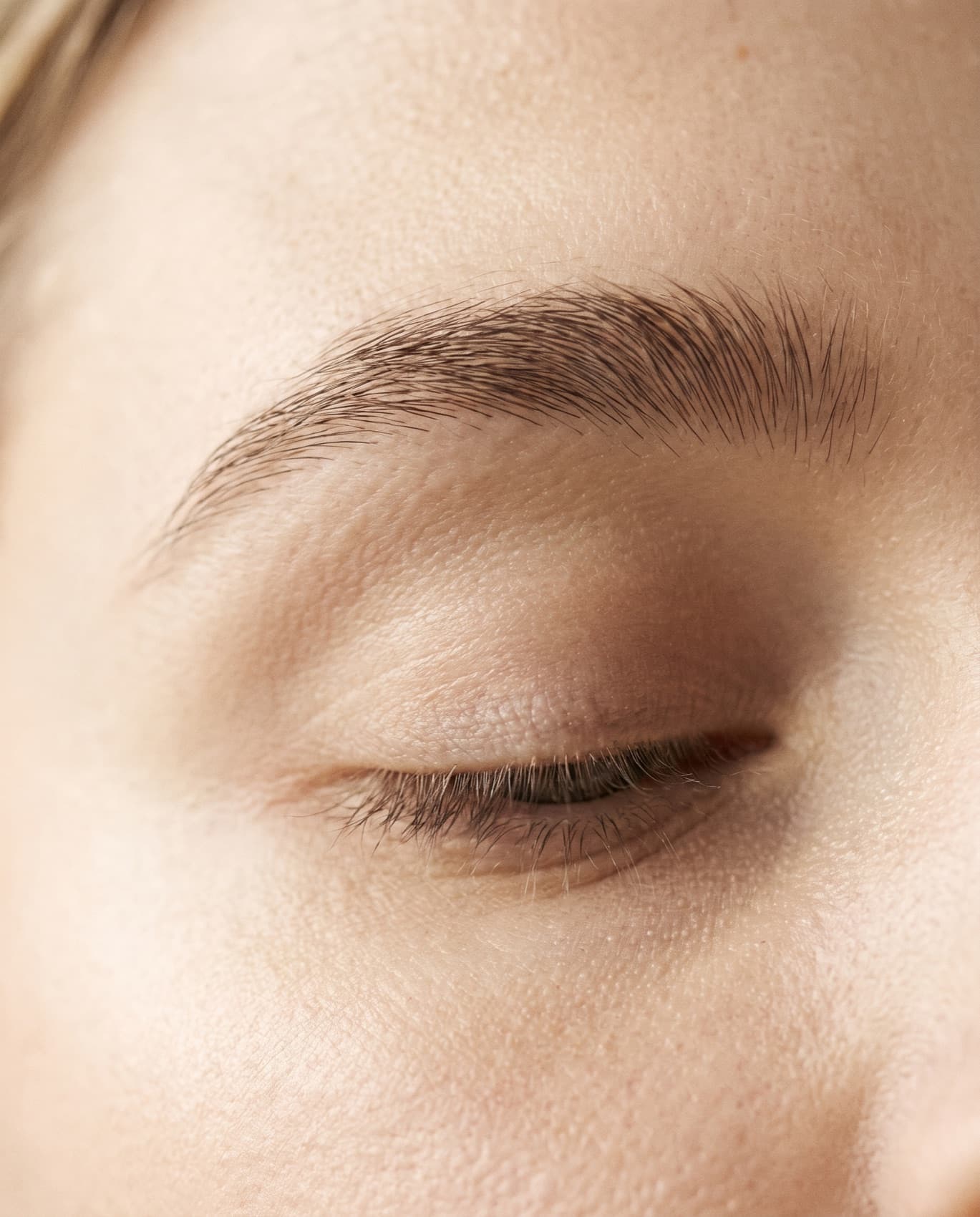 Close-up of a freshly shaped natural eyebrow above a softly closed eye, showing individual brow hairs and natural skin texture.