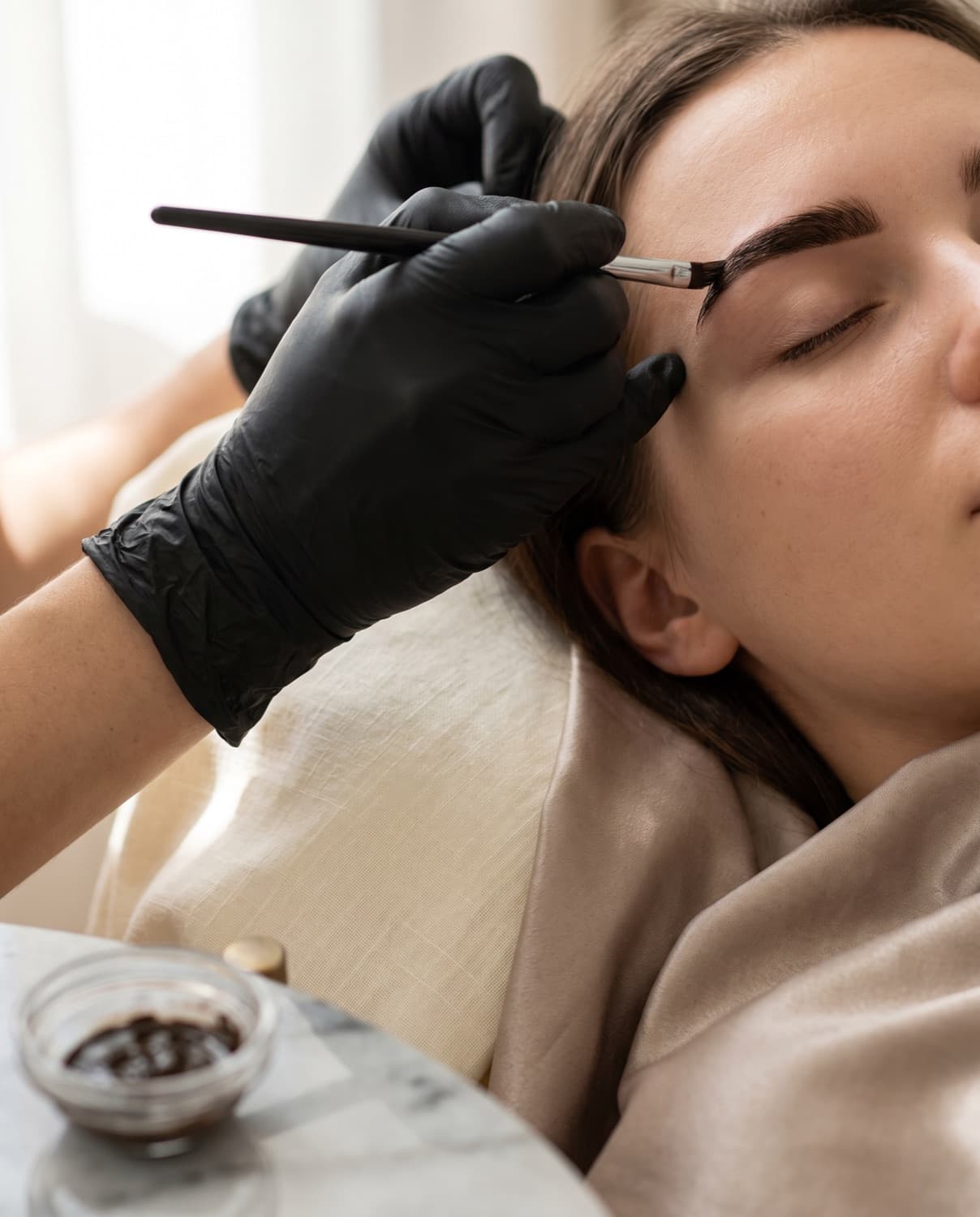 A gloved technician applying dark brow tint to a client's eyebrow with a slim angled brush.